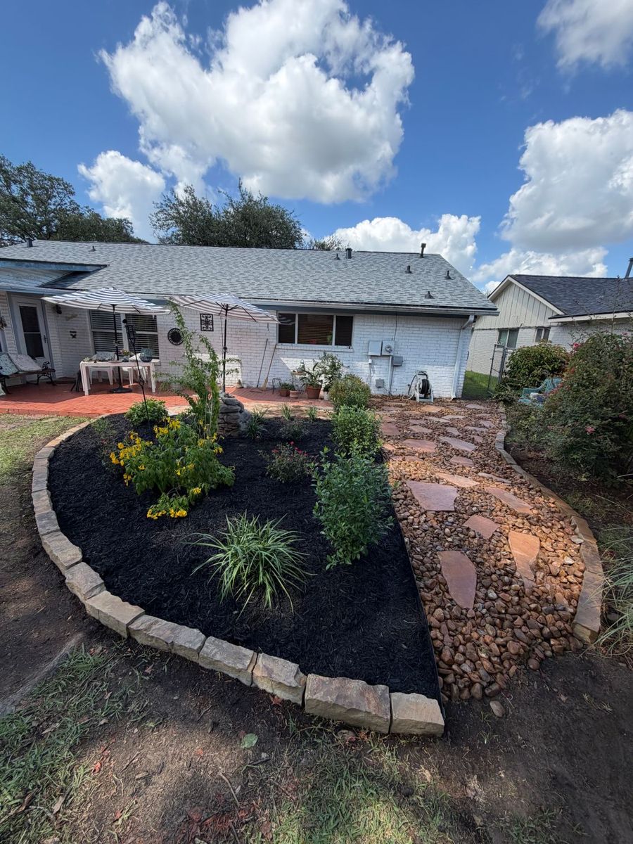 Backyard bed with a stone border, black mulch and flowering plants, next to a flagstone path set in river rock