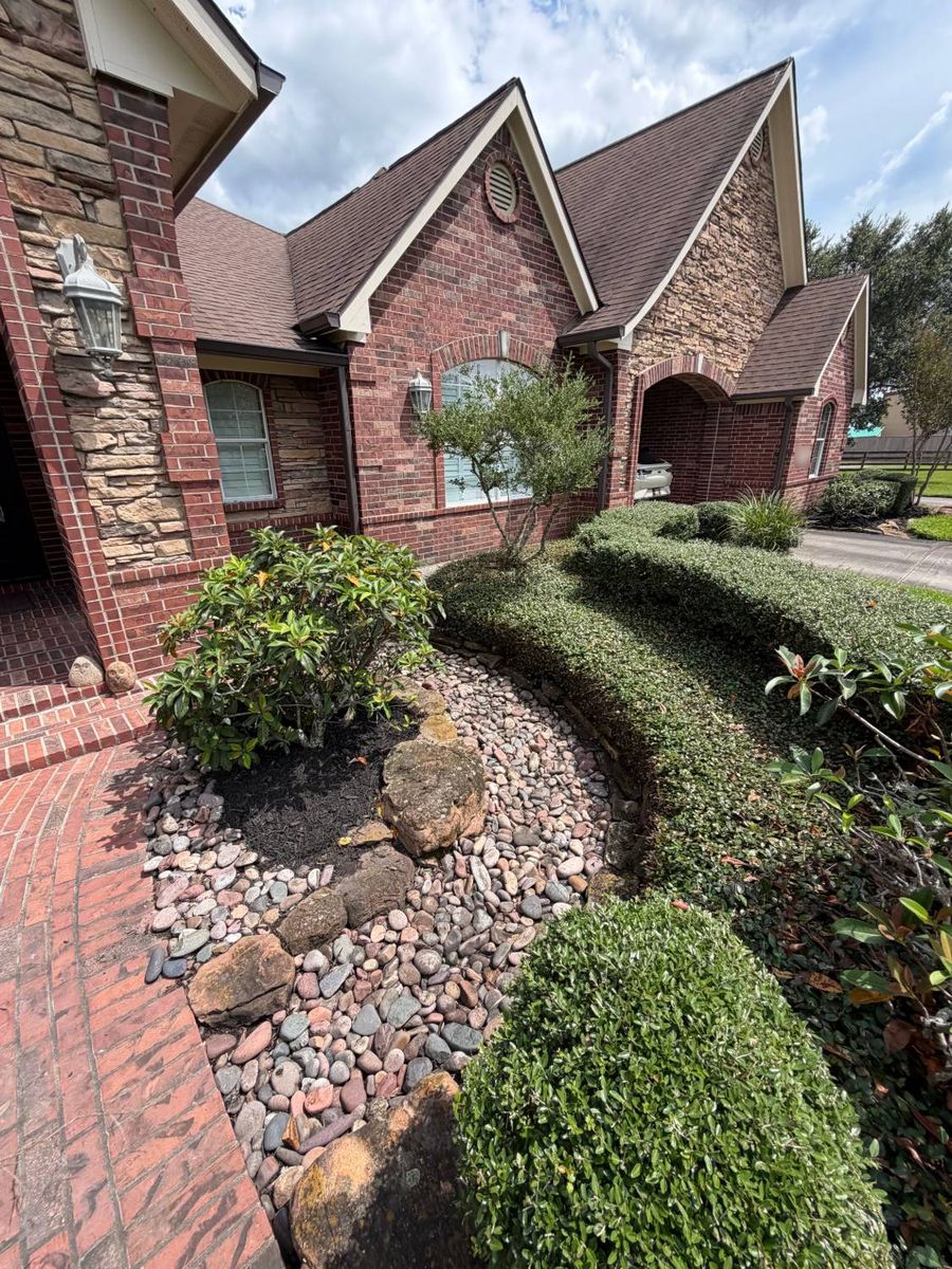 Entry bed of river rock and boulders around a shrub, framed by a curved trimmed hedge beside a brick walkway