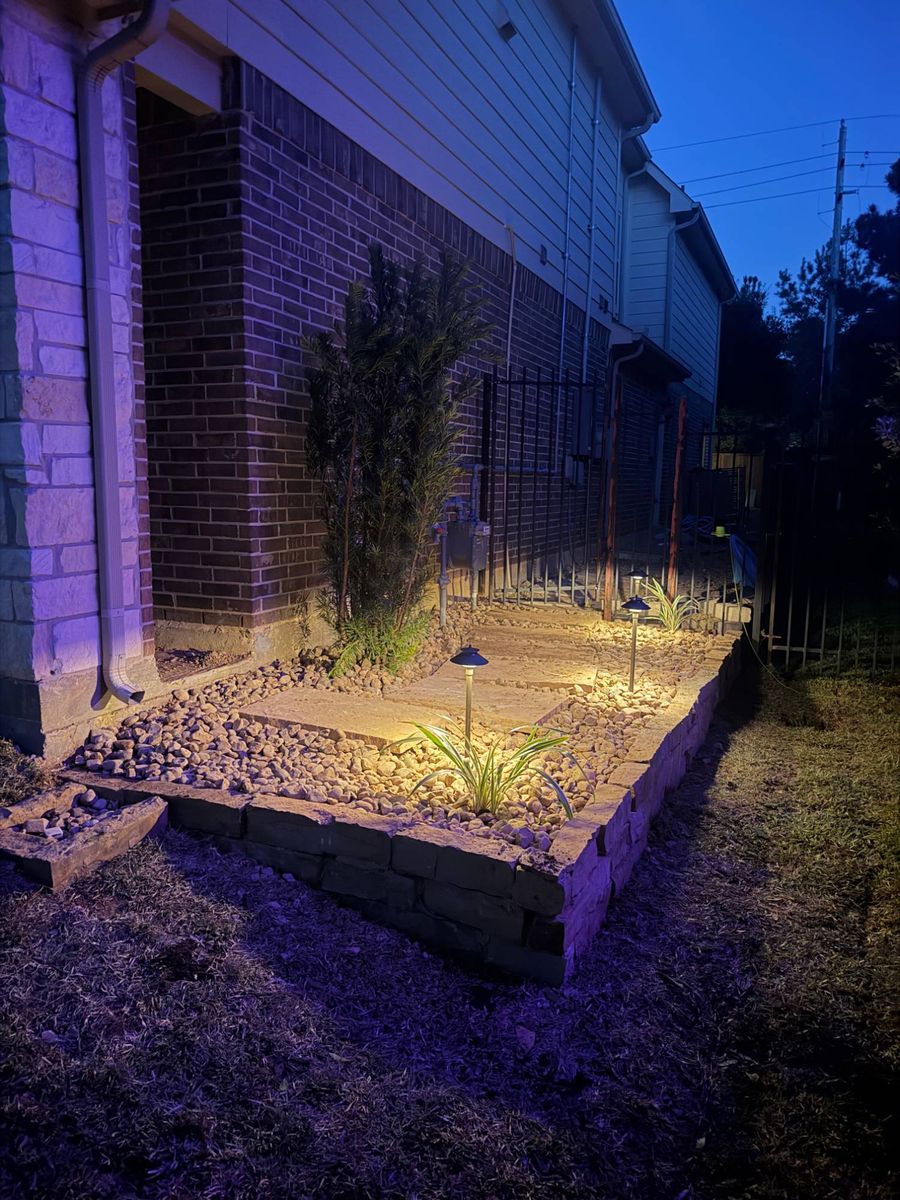 Path lights glowing in a raised stone-bordered bed of river rock and flagstone along the side of a brick house at dusk
