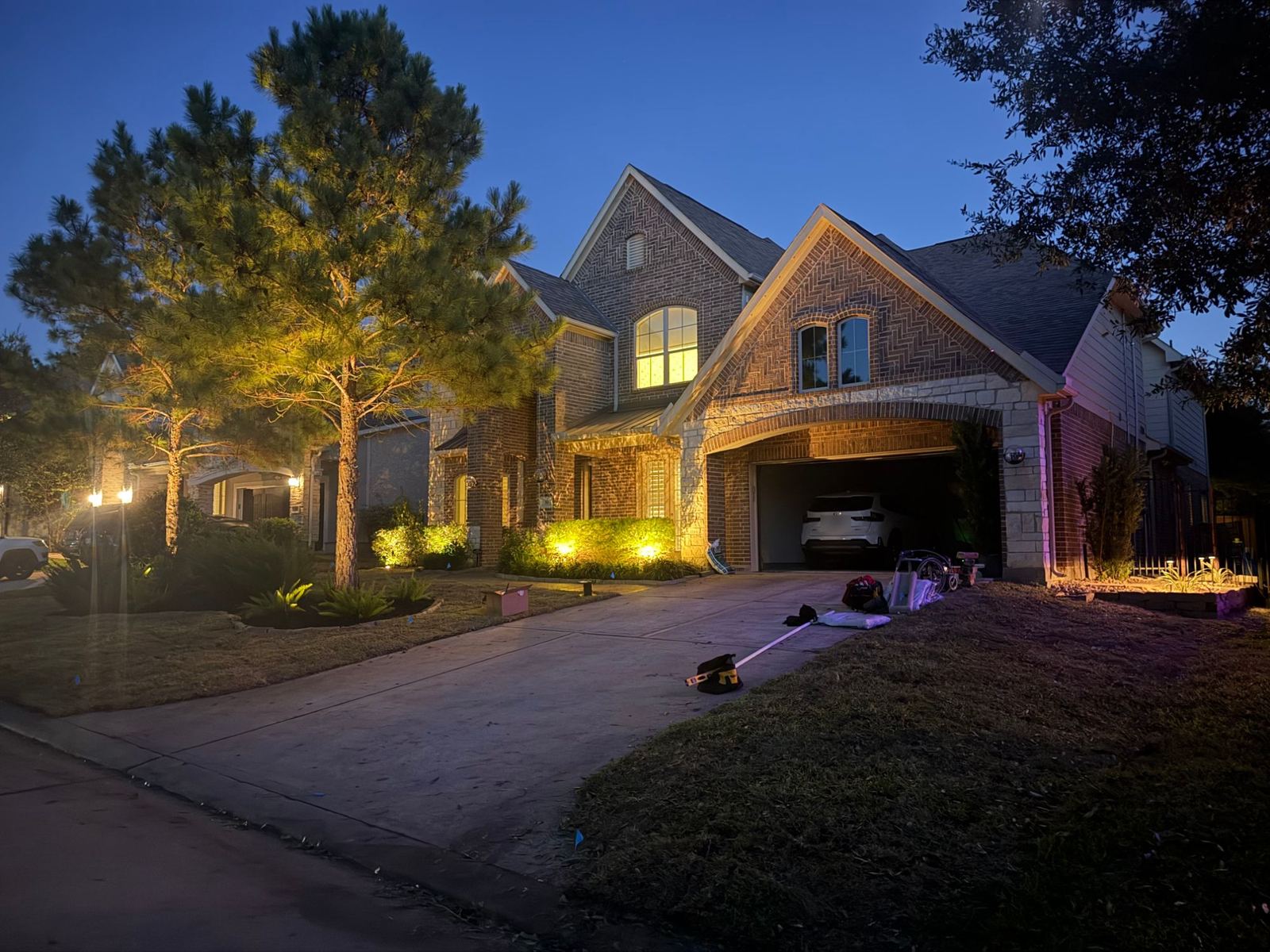 Two-story brick home at dusk with uplit pine trees, lit shrub beds along the front and a lit side bed